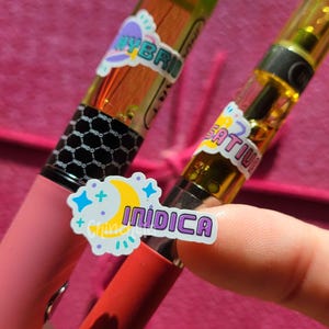 Puede incluir: Dos cartuchos de vapeo con aceite amarillo claro, uno con boquilla verde y etiqueta Hybrid, el otro con etiqueta Sativa. En primer plano se sostiene una pegatina separada con una luna creciente amarilla y el texto Indica.