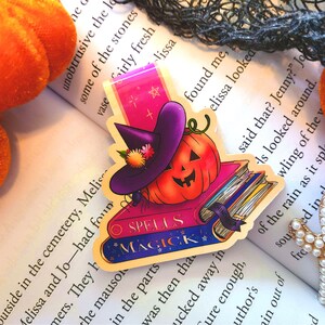 Puede incluir: Un marcapáginas con temática de Halloween con una calabaza con un sombrero de bruja, una pila de libros y las palabras "Spells Magick".