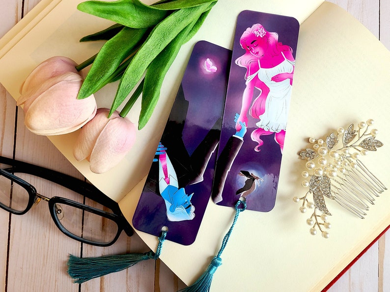 May include: Two colorful bookmarks with a dark purple background. One bookmark features a woman with pink hair and a white dress, the other features a man with blue hair and a black suit. Both bookmarks have a teal tassel.