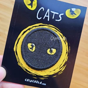 May include: A circular black embroidered patch featuring two yellow cat eyes, mounted on a black rectangular backing card. The card displays the word CATS in white text at the top and the website address CHINCHELA.com in white text at the bottom.