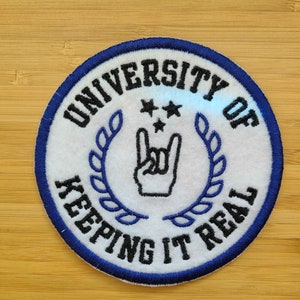 以下が含まれることがあります： 青い縁取りのある白い刺繍パッチ。パッチには、ロックンロールの手のジェスチャーをしている手の白黒のイラストの周りに、黒い文字で「University of Keeping It Real」というテキストが書かれています。手の真上に小さな星が2つあります。