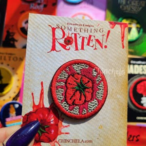 May include: A circular embroidered patch featuring a red tomato design with a green stem, set against a textured background. The patch is mounted on a display card with the text Broadway Badges SOMETHING ROTTEN! and a red tomato graphic.