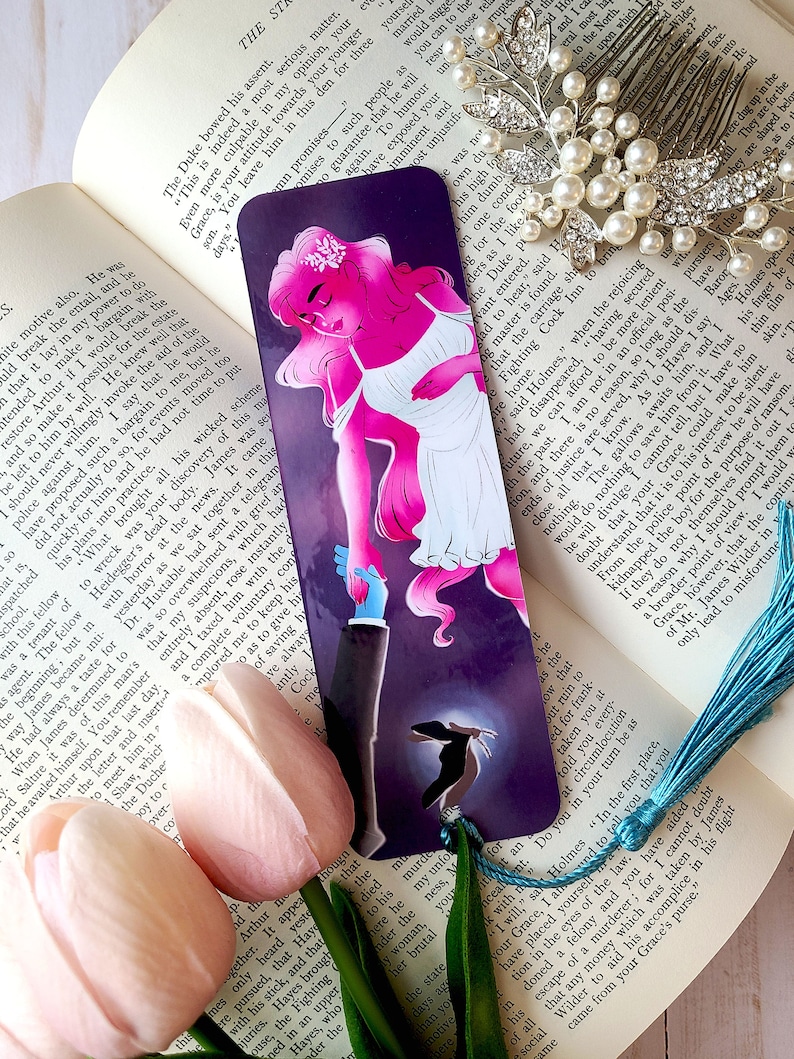 May include: A bookmark featuring a pink and white illustration of a woman with a crown of flowers and a man in a suit. The woman is holding the man's hand and they are standing on a purple background with a silhouette of a woman in the background. The bookmark has the text "The Stranger" at the top.