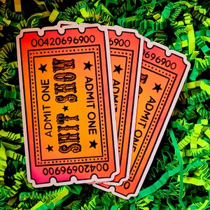 May include: Three orange, pink, and yellow ticket-shaped stickers with the text "SHIT SHOW" and "ADMIT ONE" printed on them. The stickers are on a green and yellow shredded paper background.