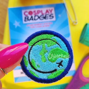May include: A circular embroidered patch featuring a stylized globe with light blue oceans and green landmasses, accented by a black curved line resembling a smile and a small black airplane icon, all bordered by a thick dark blue stitched edge.