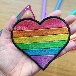 Pode incluir: Um emblema bordado em forma de coração com listras horizontais em vermelho, laranja, amarelo, verde, azul e roxo, com uma borda preta espessa. A palavra Chinchela está impressa em letra cursiva branca no lado esquerdo do emblema.