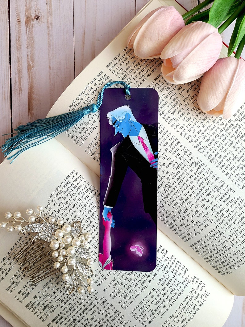 May include: A bookmark featuring a blue-skinned man in a black suit and red tie, holding the hand of a pink-skinned woman with a butterfly behind them. The bookmark has a teal tassel.