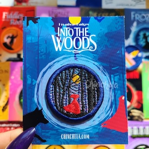 May include: A circular embroidered patch featuring a forest scene with a figure in a red cloak and a yellow moon, mounted on a blue backing card with the text INTO THE WOODS and CHINCHELA.COM printed in white, showcasing a detailed thread design.