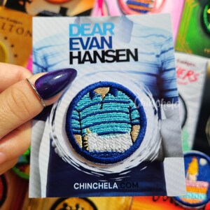 May include: A circular embroidered patch featuring a blue and white striped polo shirt with a white arm cast. The patch is mounted on a display card with the text DEAR EVAN HANSEN at the top and CHINCHELA.COM at the bottom, set against a blue background.
