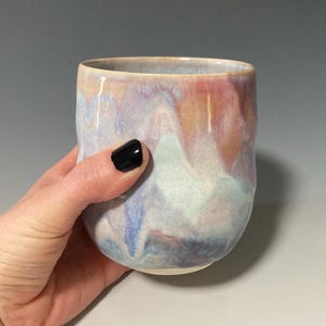May include: A handcrafted ceramic tumbler with a distinctive, colourful glaze. The cup displays a mix of pastel shades, such as pink, blue, and cream, producing a marbled appearance. The rounded form and smooth surface suggest a comfortable grip, ideal for drinks.
