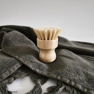 May include: A wooden brush with natural bristles, resting on a dark green towel with tassels. The brush is designed for cleaning dishes or other surfaces.