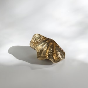 May include: A gold-coloured metal seashell-shaped dish. The dish is smooth and has a wavy texture.
