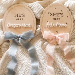 Puede incluir: Dos letreros ovalados de madera con los textos "HE'S HERE" y "SHE'S HERE" en letra cursiva. El primer letrero tiene el nombre "Gregory Mark" y una cinta azul. El segundo letrero tiene el nombre "Ayla Marie" y una cinta rosa. Ambos letreros tienen un borde festoneado.