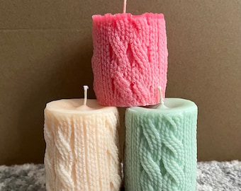 May include: Three pillar candles with a knitted texture. The candles are pink, cream, and green.