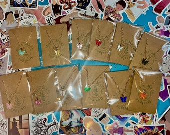 May include: A collection of 10 necklaces with butterfly charms. The necklaces are packaged on brown cards with the text "Mara Made Designs" printed on them. The butterfly charms are in various colors, including green, blue, red, orange, and white.