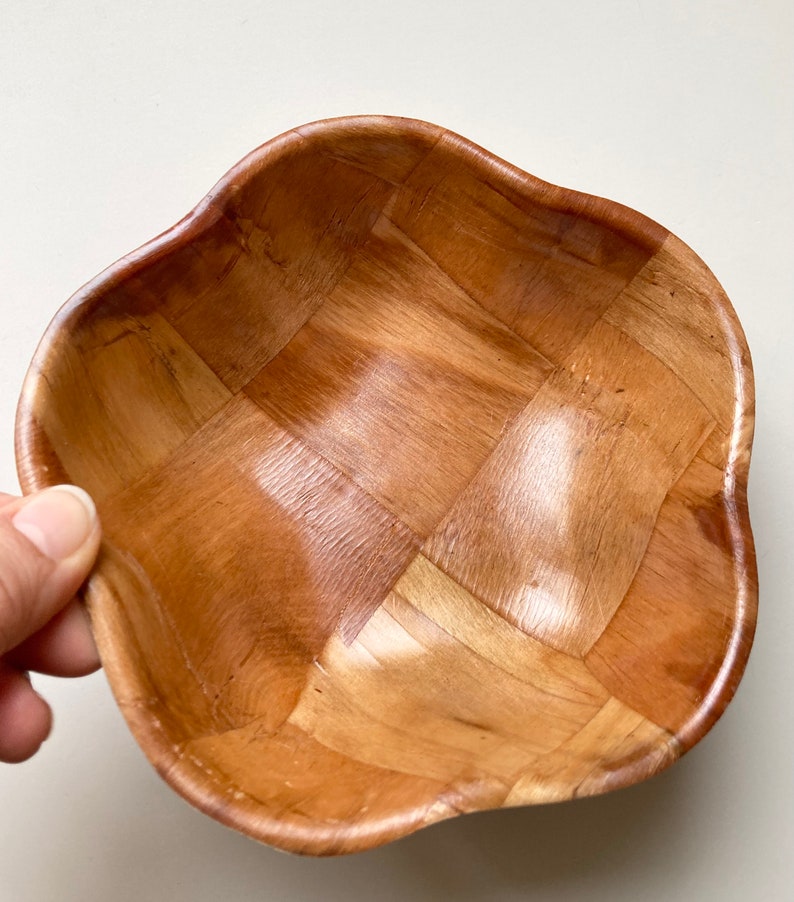 May include: A woven wooden bowl with a wavy edge. The bowl is made of light brown wood and has a square pattern.