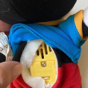 May include: A plush Mickey Mouse doll with a black head, blue jacket, and red pants. The doll's back is open, revealing a yellow sound module with a screw and the text "SMART TOY". The doll's tag is visible.