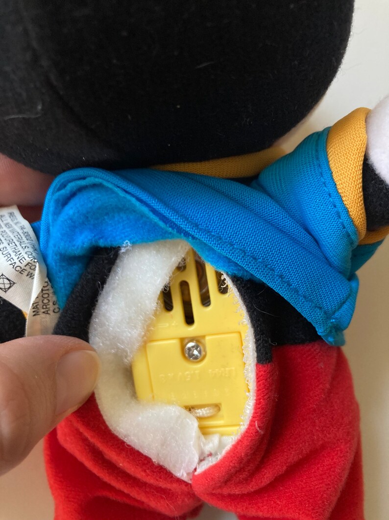 May include: A plush Mickey Mouse doll with a black head, blue jacket, and red pants. The doll's back is open, revealing a yellow sound module with a screw and the text "SMART TOY". The doll's tag is visible.