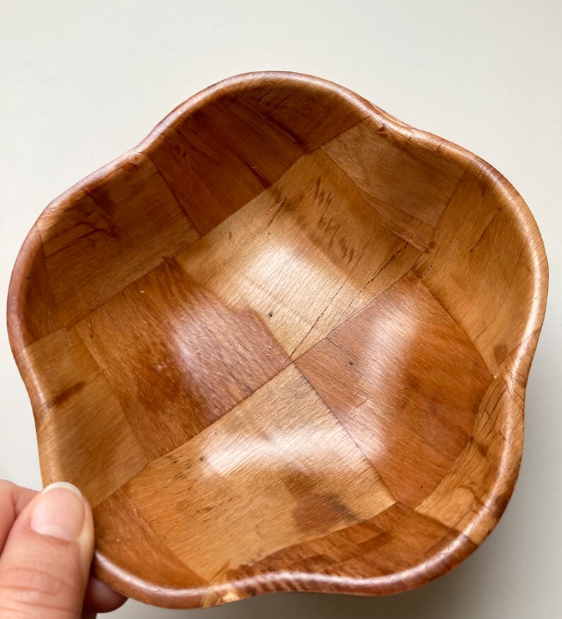 May include: A woven wooden bowl with a light brown color and a wavy rim. The bowl is made of thin strips of wood that are woven together to create a checkered pattern.