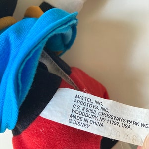 May include: A close-up of a Mickey Mouse plush toy. The toy has a red bottom, black shorts, a blue jacket, and white gloves. The tag reads "MATTEL, INC. ARCOTOYS, INC. MADE IN CHINA &copy; DISNEY."