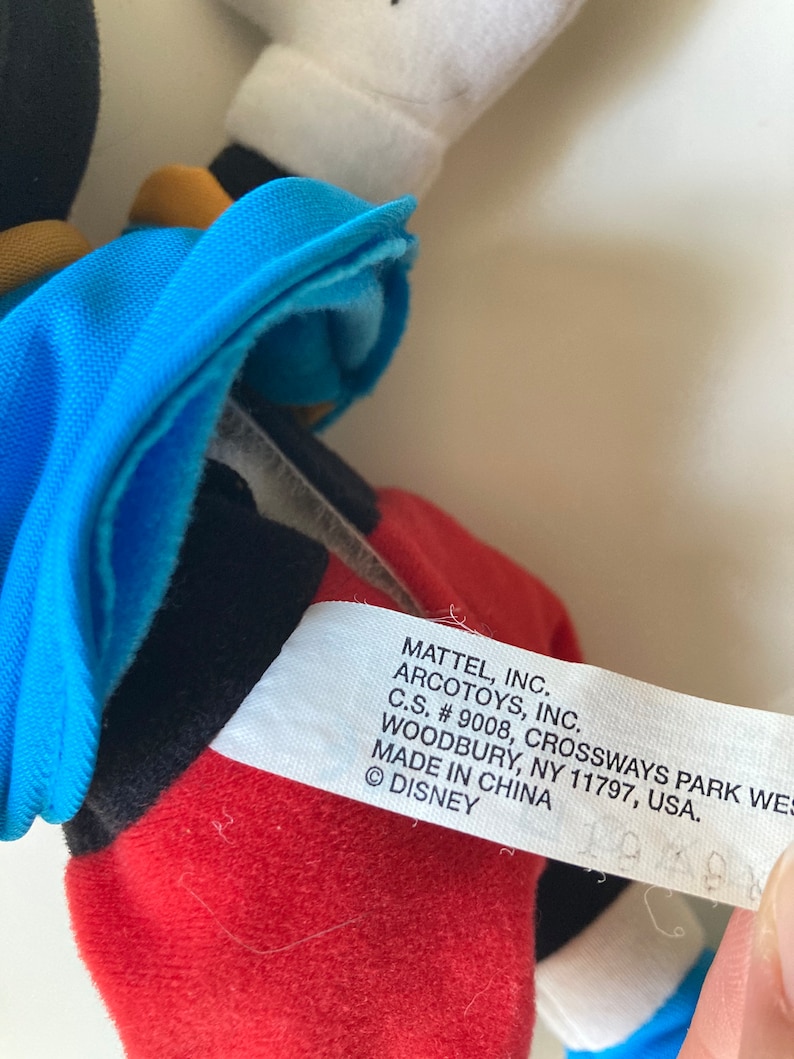 May include: A close-up of a Mickey Mouse plush toy. The toy has a red bottom, black shorts, a blue jacket, and white gloves. The tag reads "MATTEL, INC. ARCOTOYS, INC. MADE IN CHINA &copy; DISNEY."