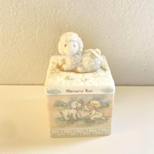May include: A white ceramic memory box with a lamb figurine on top. The box has a colorful design of a child and a lamb with the words "Memory Box" printed on the front.