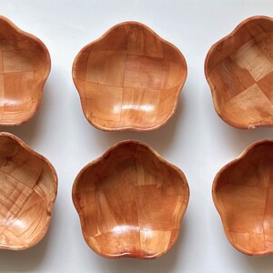 May include: Six small, wooden bowls in a light brown color. The bowls are shaped like a flower with five petals and have a woven pattern.