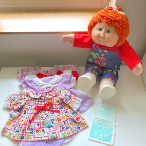 May include: Cabbage Patch Kid doll with red yarn hair wearing a red shirt and blue overalls with a floral pattern. The doll is sitting on a white surface with a stack of four doll dresses in various colors and patterns. A birth certificate for the doll is visible in the lower right corner of the image.