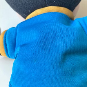 May include: A plush toy featuring a black head, a blue shirt with yellow trim, and red shorts. The toy's back is visible, showcasing the texture of the blue fabric and the stitching details.