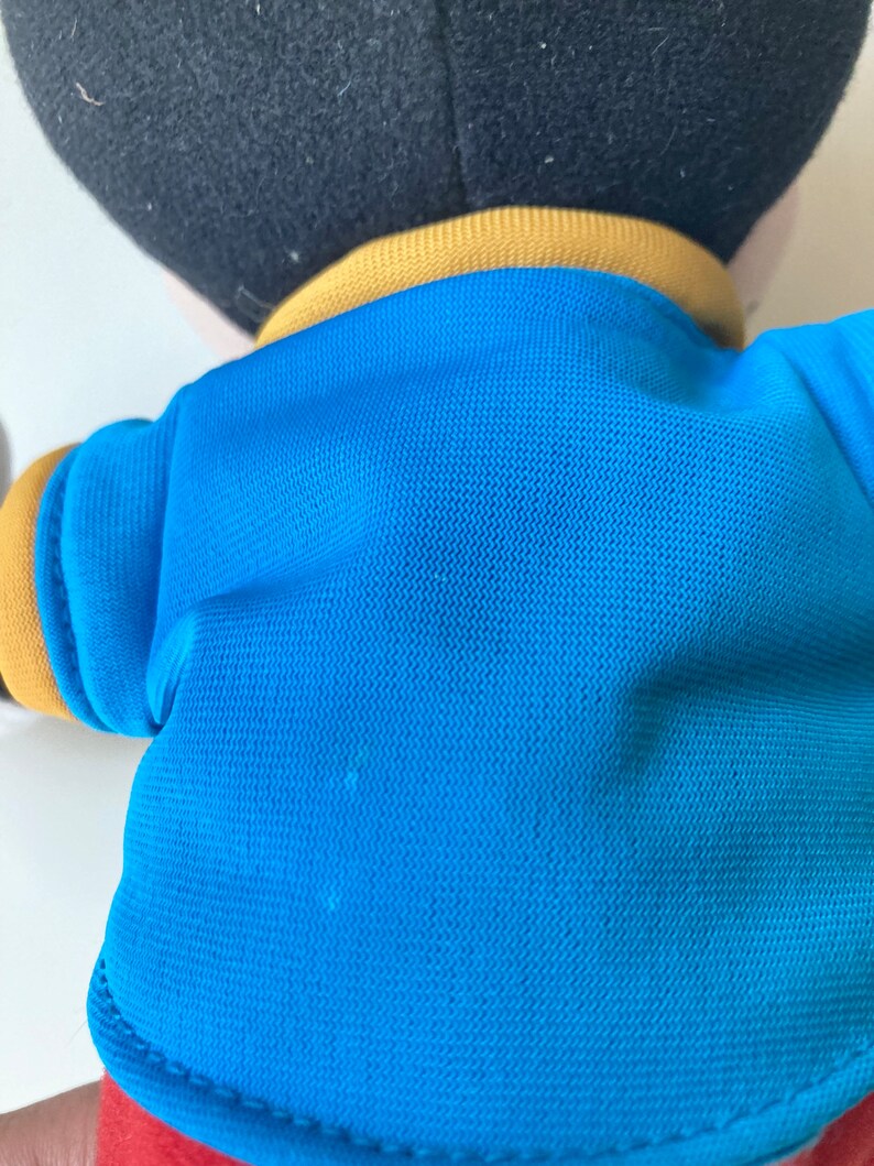 May include: A plush toy featuring a black head, a blue shirt with yellow trim, and red shorts. The toy's back is visible, showcasing the texture of the blue fabric and the stitching details.