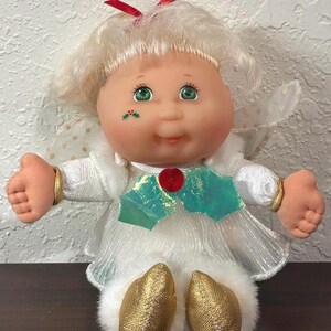 May include: A doll with blonde hair, green eyes, and a red bow on top. It wears a white dress with a red button, green holly leaf accents, and gold shoes. A small holly leaf graphic is on its cheek. It has white wings with gold dots.