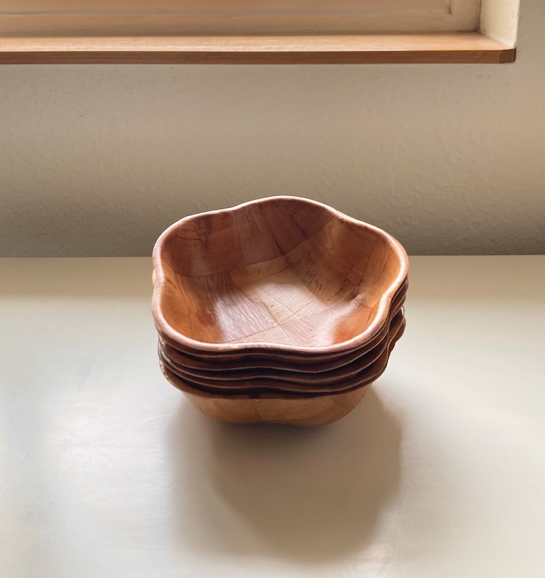 May include: A set of four wooden bowls stacked on top of each other. The bowls are a light brown color and have a wavy, flower-like shape.