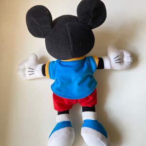 May include: A Mickey Mouse plush toy, viewed from the back, with black ears and head, a blue shirt with yellow trim, red shorts, white gloves, and blue and white shoes. The toy's arms are outstretched.