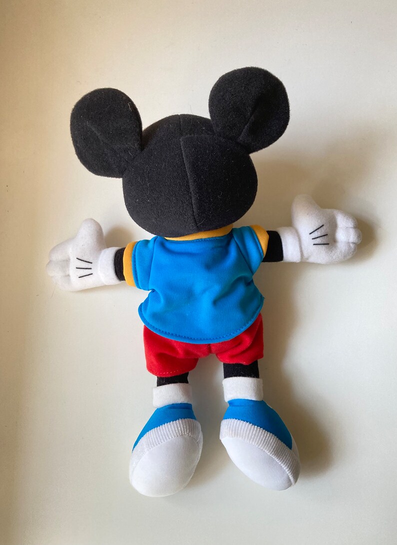 May include: A Mickey Mouse plush toy, viewed from the back, with black ears and head, a blue shirt with yellow trim, red shorts, white gloves, and blue and white shoes. The toy's arms are outstretched.