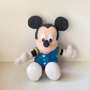 May include: A plush Mickey Mouse doll with black ears, a tan face, and large black eyes. He wears a blue jacket with yellow trim, red shorts, and white gloves and shoes. The doll is sitting with its arms outstretched.