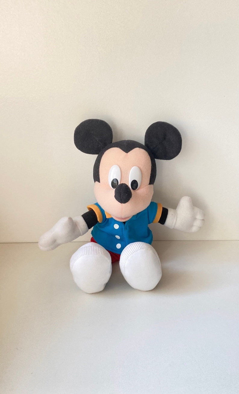 May include: A plush Mickey Mouse doll with black ears, a tan face, and large black eyes. He wears a blue jacket with yellow trim, red shorts, and white gloves and shoes. The doll is sitting with its arms outstretched.