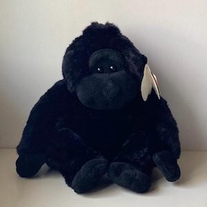 May include: A plush, black gorilla toy with a soft, furry texture. The gorilla is sitting with its arms and legs extended. It has a black face and small, dark eyes. A white tag is attached to its ear.