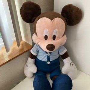 May include: A large plush Mickey Mouse toy wearing a blue shirt and blue pants. The toy has brown ears, a pink face, and black eyes.