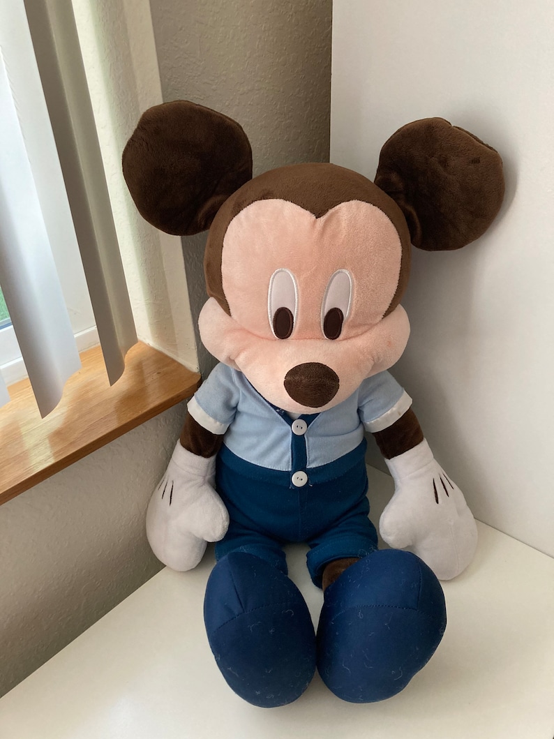 May include: A large plush Mickey Mouse toy wearing a blue shirt and blue pants. The toy has brown ears, a pink face, and black eyes.