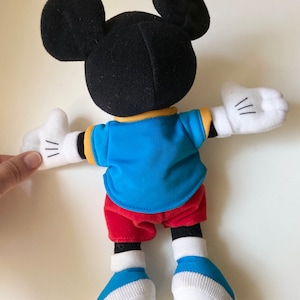 May include: A plush Mickey Mouse doll, viewed from the back, with black ears and head, wearing a blue shirt, red shorts, and white gloves and shoes. The doll has a yellow collar and white cuffs. The doll's arms are outstretched.