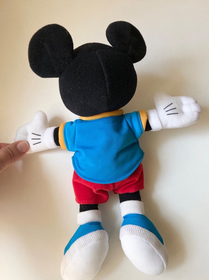 May include: A plush Mickey Mouse doll, viewed from the back, with black ears and head, wearing a blue shirt, red shorts, and white gloves and shoes. The doll has a yellow collar and white cuffs. The doll's arms are outstretched.