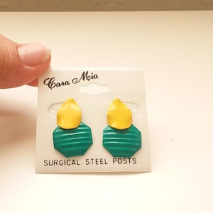 May include: A pair of green and yellow geometric earrings with surgical steel posts. The earrings are on a white card with the text "Cara Mia" and "Surgical Steel Posts".