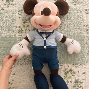 May include: A plush Mickey Mouse toy wearing a blue shirt with a bow tie and blue pants. The toy has brown ears, a brown nose, and a big smile.