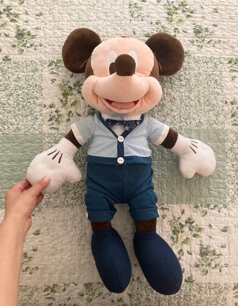 May include: A plush Mickey Mouse toy wearing a blue shirt with a bow tie and blue pants. The toy has brown ears, a brown nose, and a big smile.