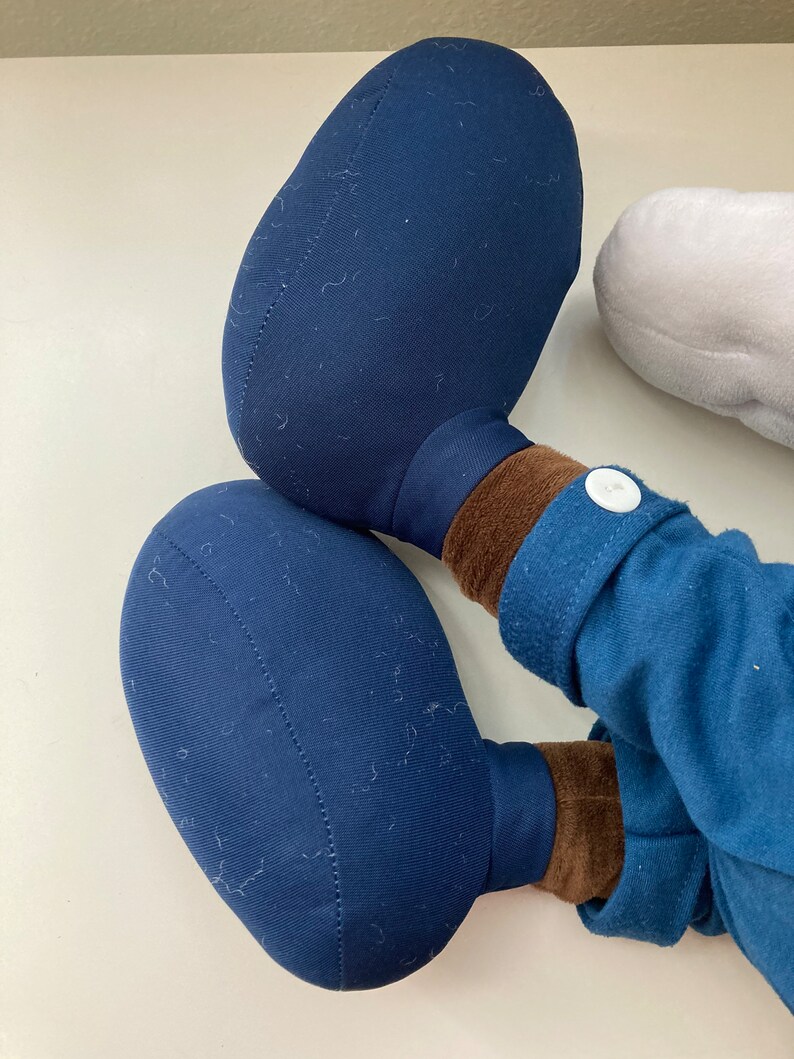 May include: A plush toy with blue fabric and brown fabric accents. The toy has two large, round, blue feet.