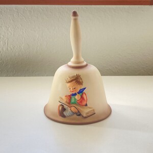 May include: A white ceramic bell with a brown handle and a painted image of a boy reading a book. The boy is wearing a green shirt, blue pants, and a red jacket.