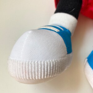 May include: Close-up of a white and blue sneaker on a plush toy. The sneaker has a white toe cap with vertical ridges, white laces, and blue accents. The toy's black leg and red clothing are visible.