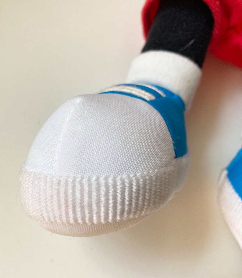 May include: Close-up of a white and blue sneaker on a plush toy. The sneaker has a white toe cap with vertical ridges, white laces, and blue accents. The toy's black leg and red clothing are visible.