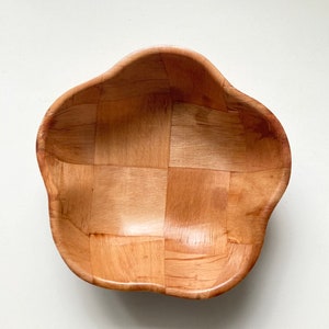 May include: A wooden bowl with a unique, wavy, flower-shaped rim. The bowl is made of woven wood panels and has a natural wood color.
