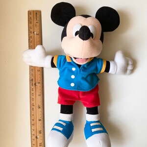 May include: A Mickey Mouse plush toy, wearing a blue jacket, red shorts, and blue shoes. The toy is approximately 12 inches tall, with a ruler placed next to it for scale. The classic character has black ears and white gloves.